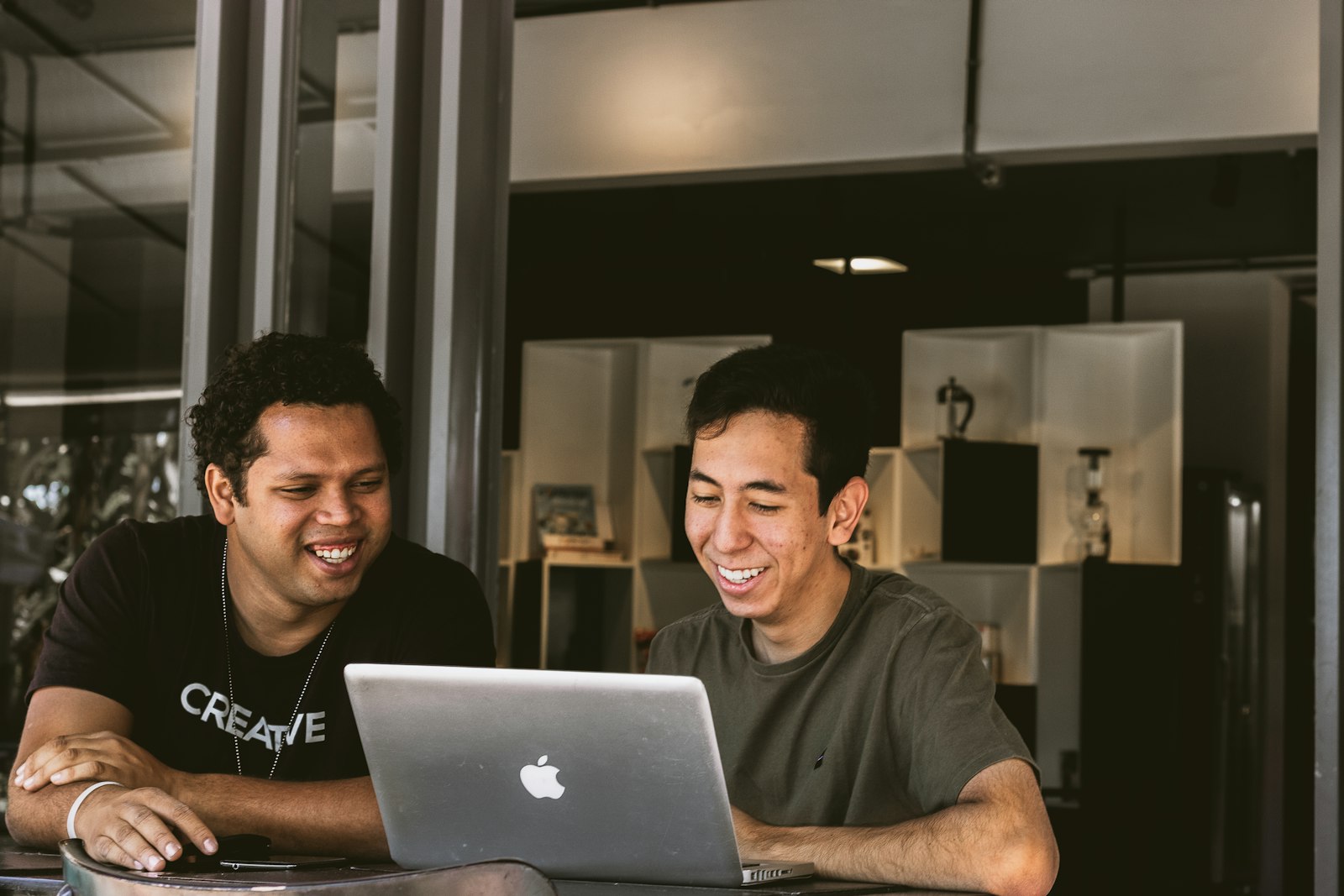 Two colleagues laughing together over one laptop at an office table.