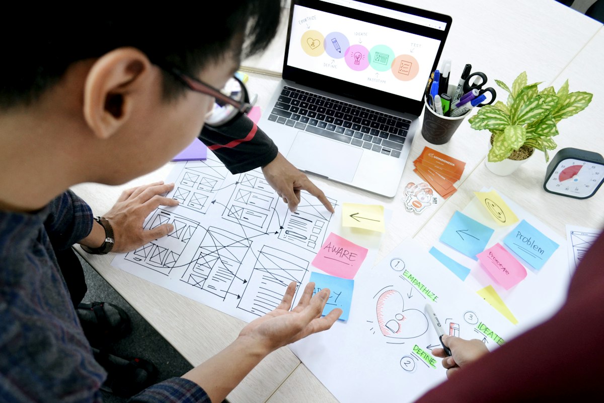 A designer talking through paper wireframes and sticky notes, a laptop open beside the drawings.